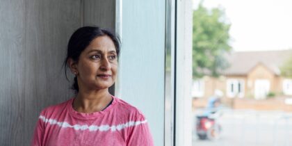 South Asian woman wearing a pink and white stripped shirt, looking into the distance from her window.