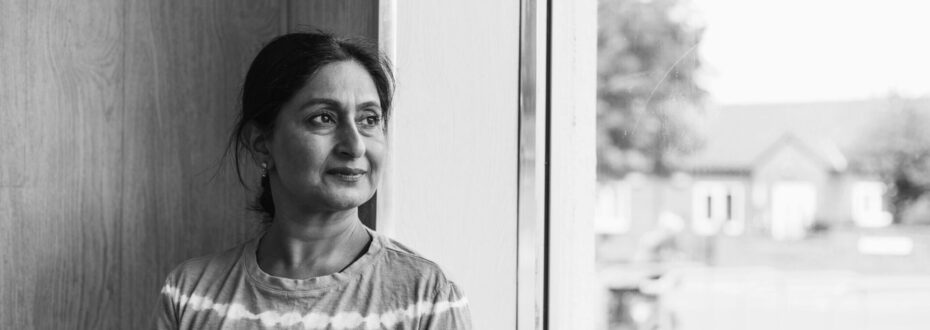 South Asian woman wearing a pink and white stripped shirt, looking into the distance from her window, in black and white.