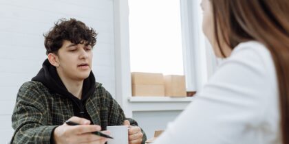 Young white boy with brunette hair, wearing a black hoodie and holding a pen in his hand, sits opposite a young white brunette woman who is listening to him speak.