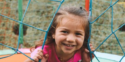 Young girl wearing a pink Mickey Mouse top smiles at camera holding onto ropes in a playground.