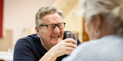 Old white man wearing glasses, holding a glass of water chuckles across the table from his friend.