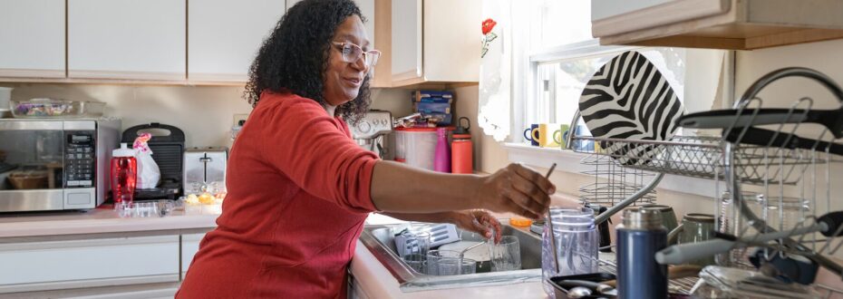 Black woman wearing red jumper doing the washing up at home.