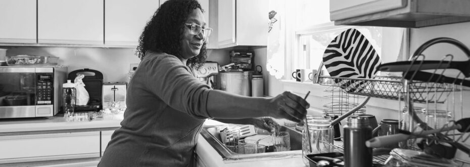 Black woman wearing red jumper doing the washing up at home, black and white image.