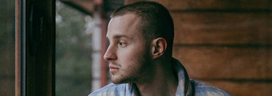 Side profile of a young white man, wearing a checkered flannel looks out of a window.