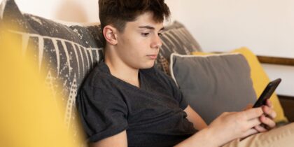 Young white boy sitting on a couch, holding a smartphone in his hand, focusing on the device with a slight frown on his face.