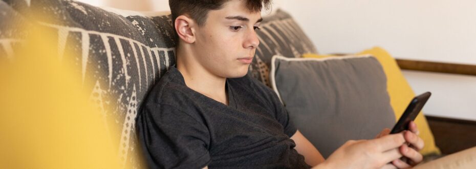 Young white boy sitting on a couch, holding a smartphone in his hand, focusing on the device with a slight frown on his face.