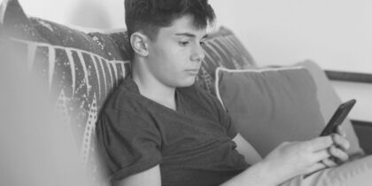Young white boy sitting on a couch, holding a smartphone in his hand, focusing on the device with a slight frown on his face. Photo is in Black and White.