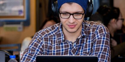 A white man wearing glasses, a blue beanie, and a plaid shirt is smiling and working on a laptop with headphones on in a workspace setting.