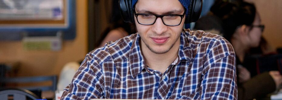 A white man wearing glasses, a blue beanie, and a plaid shirt is smiling and working on a laptop with headphones on in a workspace setting.