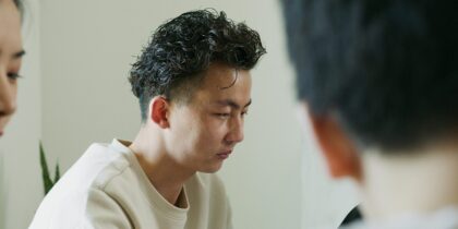 A young asian man with curly hair wearing a white sweater, sits on a chair with a look that suggests he's disappointed or unsatisfied.