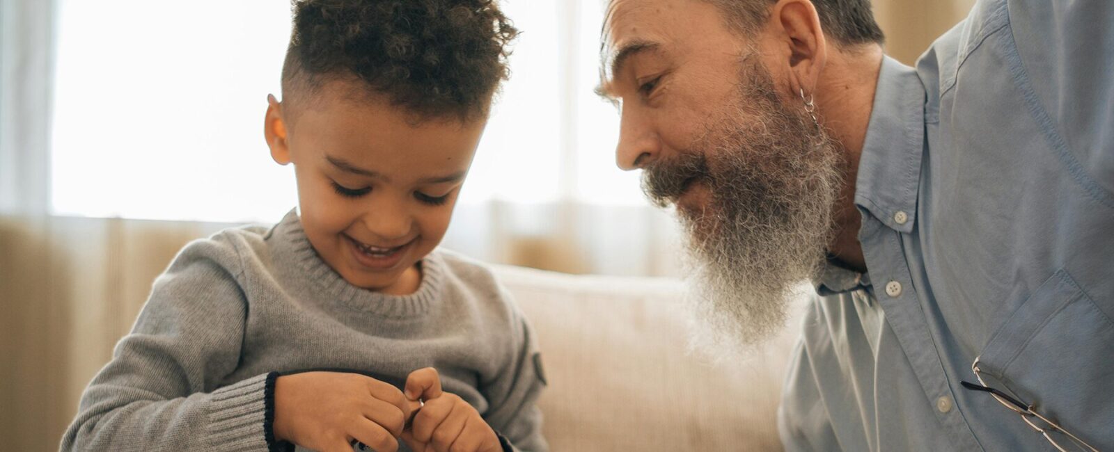 A young child, with curly dark hair, is seated on the couch next to his grandfather, who has a long beard and is wearing a gray sweater. His grandfather watches over him as he plays with his toy cars.
