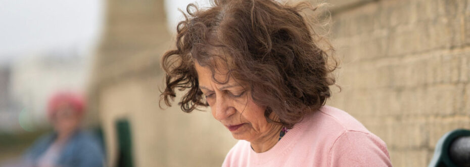 Elderly white woman with curly brown hair, wearing a light pink sweater, looking down with a pensive expression on her face.