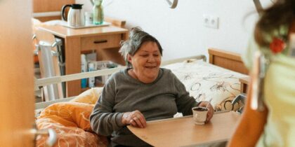 Older white women in care is sat on her bed smiling and holding a cup of tea, and her carer is smiling too.