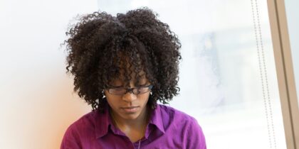 Young black woman with dark brown, curly hair wearing glasses and a dark purple buttoned shirt and a necklace, is stood with her head down.