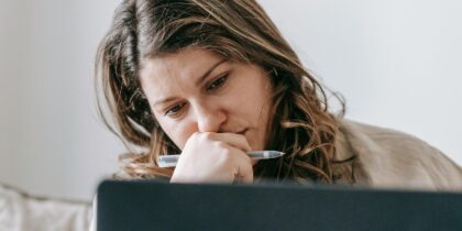 Young white woman with dark brown hair wearing a cream jumper is holding a pen in her hands over her mouth, and thinking about what she's reading on her laptop screen.