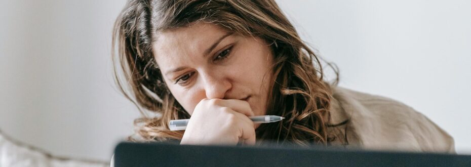 Young white woman with dark brown hair wearing a cream jumper is holding a pen in her hands over her mouth, and thinking about what she's reading on her laptop screen.