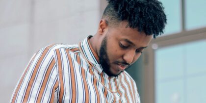 A young Black man with curly dark hair and a beard, wearing a striped button-up shirt stands with his head down with a thoughtful expression on his face.