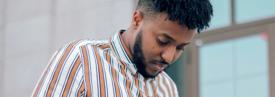 A young Black man with curly dark hair and a beard, wearing a striped button-up shirt stands with his head down with a thoughtful expression on his face.