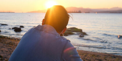 Photograph from behind of a person sitting on a beach looking towards the sun setting over the sea