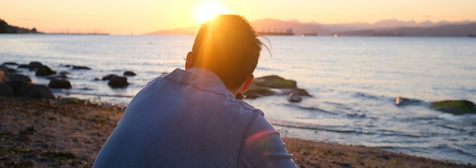 Photograph from behind of a person sitting on a beach looking towards the sun setting over the sea