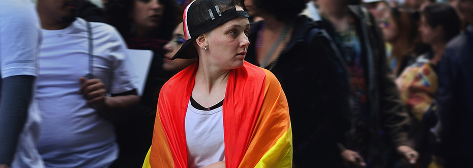 A young person draped in a rainbow flag is alone in a crowd, looking off to the right