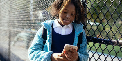 A young Black girl leaning on a chainlink fance is looking at her smartphone