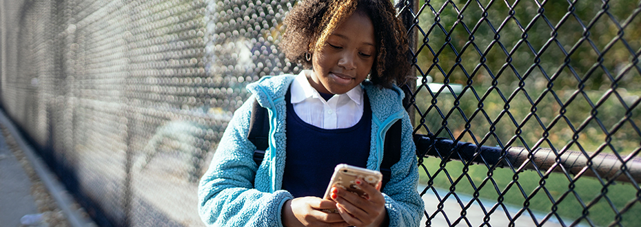 A young Black girl leaning on a chainlink fance is looking at her smartphone