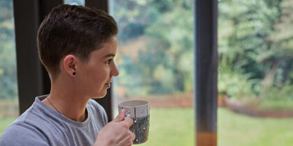A person with a coffee mug is looking out of a window towards a garden