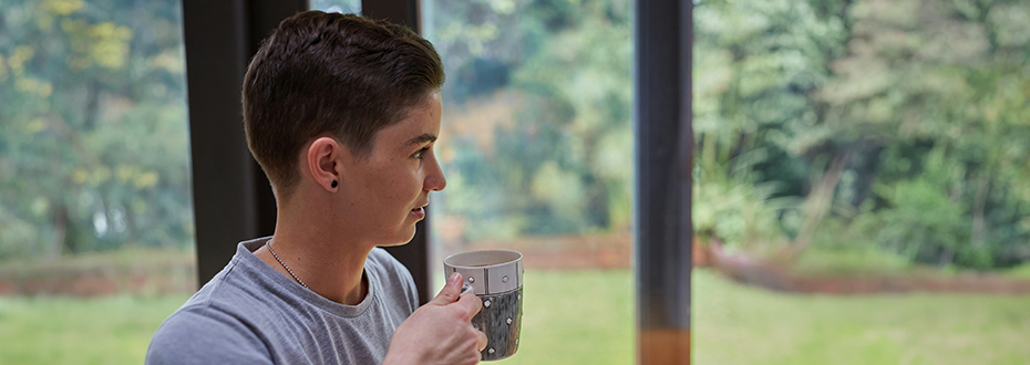 A person with a coffee mug is looking out of a window towards a garden