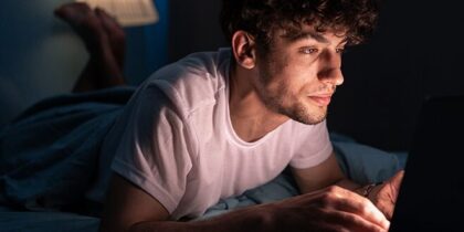 A young man lies on a bed in a darkened room, his face lit by a laptop screen