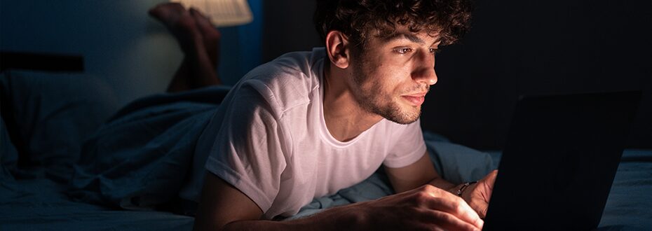 A young man lies on a bed in a darkened room, his face lit by a laptop screen