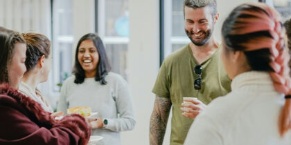A group of 6 people standing in a circle in a bright office, smiling and chatting