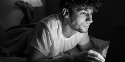 A young man lies on a bed in a darkened room, his face lit by a laptop screen