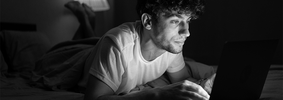 A young man lies on a bed in a darkened room, his face lit by a laptop screen