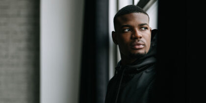 A young Black man is standing by a window looking into the distance