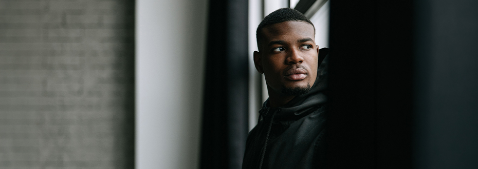 A young Black man is standing by a window looking into the distance