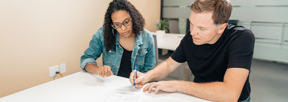 At an office desk, a woman is helping a man to fill out a form.
