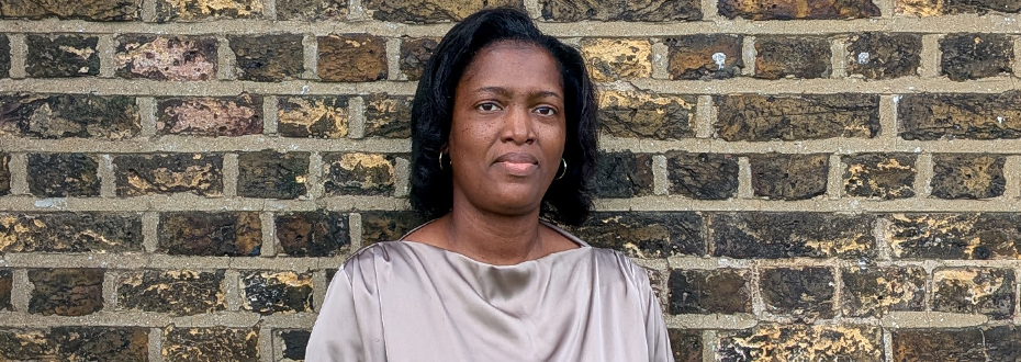 Portrait photo of a Black woman looking into the camera, standing in front of a brick wall