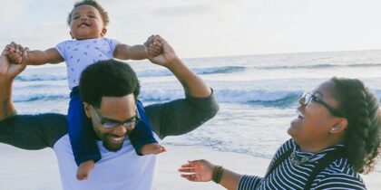Black man and woman laughing together at the seaside with baby on the shoulders of the man