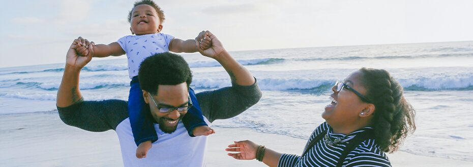 Black man and woman laughing together at the seaside with baby on the shoulders of the man