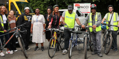 A group of 11 mostly Black folk standing in a row with bicycles and banner in the background reading A.C.C.I.