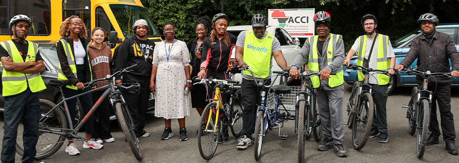 A group of 11 mostly Black folk standing in a row with bicycles and banner in the background reading A.C.C.I.