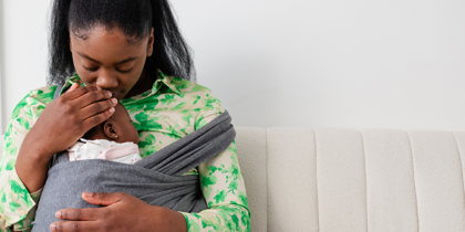A Black woman sitting on a sofa cradling her baby in a sling