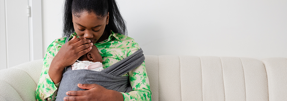 A Black woman sitting on a sofa cradling her baby in a sling