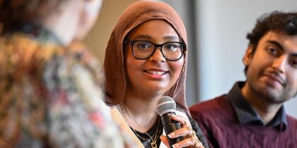 A young woman in a headscarf is speaking with a microphone while a young man looks on