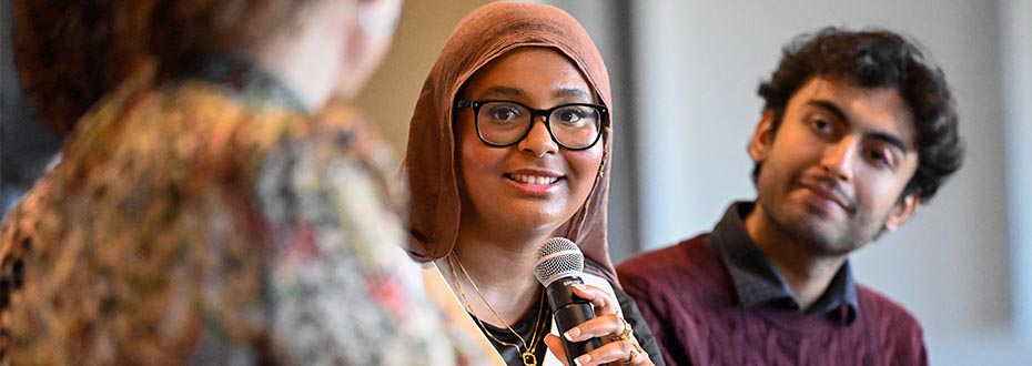 A young woman in a headscarf is speaking with a microphone while a young man looks on