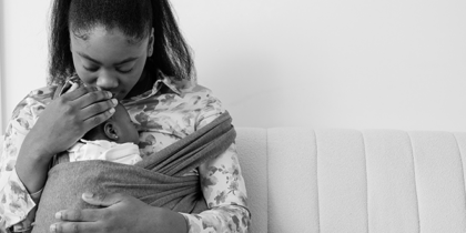 A Black woman sitting on a sofa cradling her baby in a sling