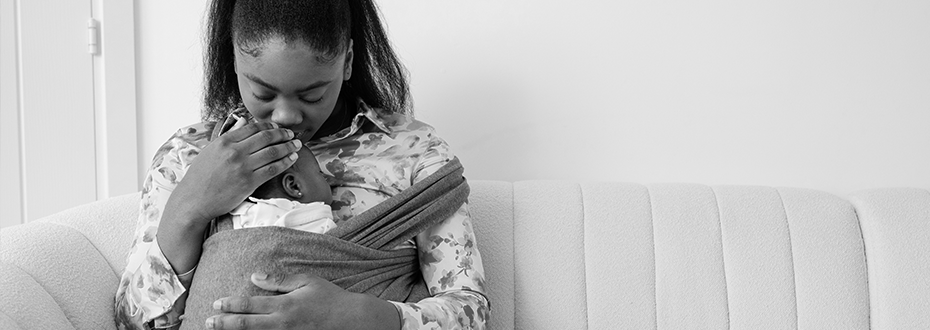 A Black woman sitting on a sofa cradling her baby in a sling