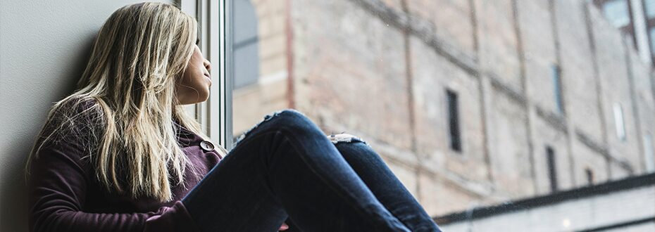 A woman sits on a windowledge looking out at buildings